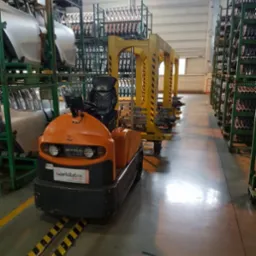 Pomarańczowy wózek transportowy z logiem Widlatex na hali produkcyjnej, przewożący elementy karoserii samochodowej na wózkach z żółtymi elementami zabezpieczającymi.