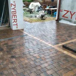 Ułożona, mokra kostka brukowa w odcieniach brązu i szarości z liniowym odwodnieniem, obramowana jaśniejszym pasem kostki, prowadząca do schodów i budynku z napisem 'ZYBERT'.