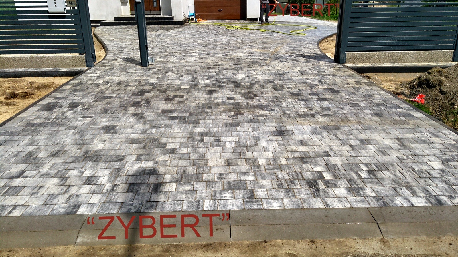 Szeroki podjazd z szarej kostki brukowej ułożonej w regularny wzór, z widocznym napisem 'ZYBERT' na obrzeżu, z bramą w tle i osobą z wężem do podlewania.