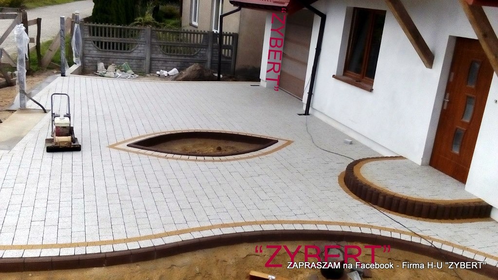 Ułożona jasnoszara kostka brukowa z dekoracyjnymi obramowaniami w kolorze brązowym i beżowym, tworząca wzór oczka wodnego i ścieżkę do drzwi wejściowych, ubijarka wibrująca na powierzchni.