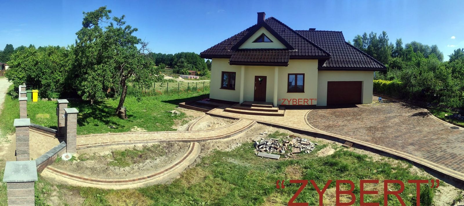 Panoramiczny widok na dom z beżową elewacją i ciemnobrązowym dachem, otoczony nowo ułożoną ścieżką z kostki brukowej, w tle zielony ogród i drzewa.