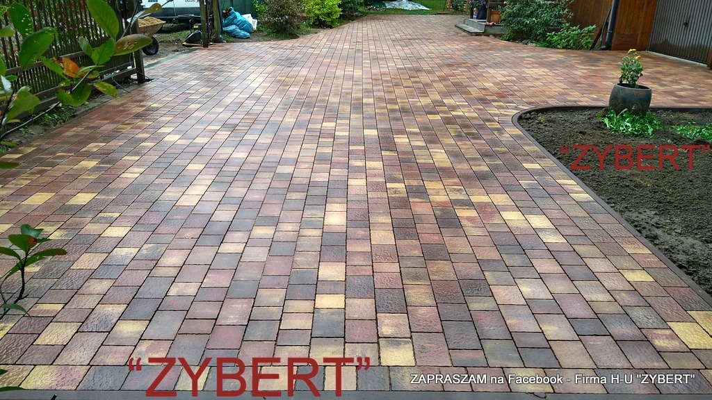 Szeroki, wybrukowany podjazd z kostki w odcieniach brązu, beżu i czerwieni, prowadzący do domu z garażem; widoczny fragment ogrodzenia i roślinność.