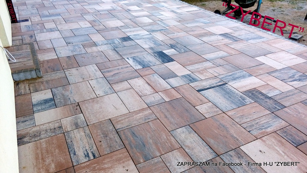 Ułożona nawierzchnia z kostki brukowej w odcieniach brązu, szarości i beżu, ułożona w nieregularny wzór. W tle widoczna betoniarka.