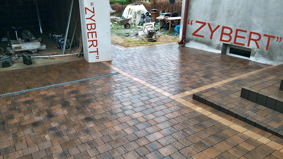 Ułożona, mokra kostka brukowa w odcieniach brązu i szarości z liniowym odwodnieniem, obramowana jaśniejszym pasem kostki, prowadząca do schodów i budynku z napisem 'ZYBERT'.