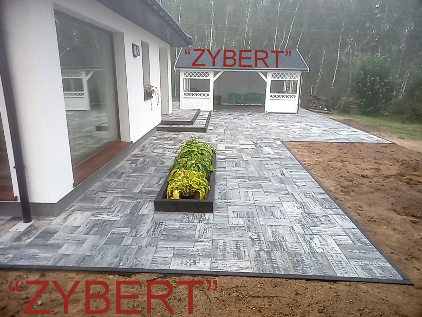 Szara kostka brukowa ułożona w nieregularny wzór przed domem z białymi ścianami i altaną w tle, w donicy posadzone rośliny