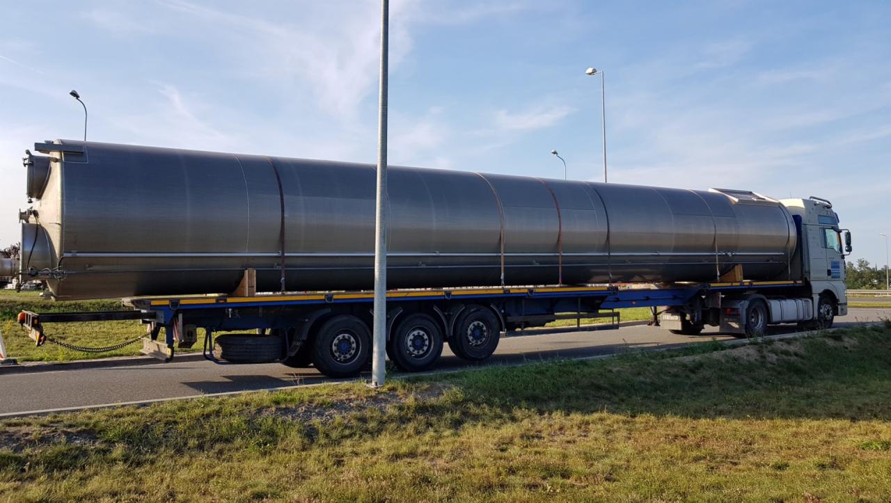 Ciężarówka z naczepą cysterną, zabezpieczona pasami, na poboczu drogi. Widok z boku, w tle błękitne niebo.