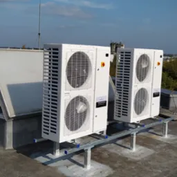 Dwa białe, bliźniacze urządzenia na dachu, prawdopodobnie klimatyzacyjne, zamontowane na metalowej konstrukcji, z widocznymi wentylatorami i elementami elektrycznymi, na tle miejskiego krajobrazu...