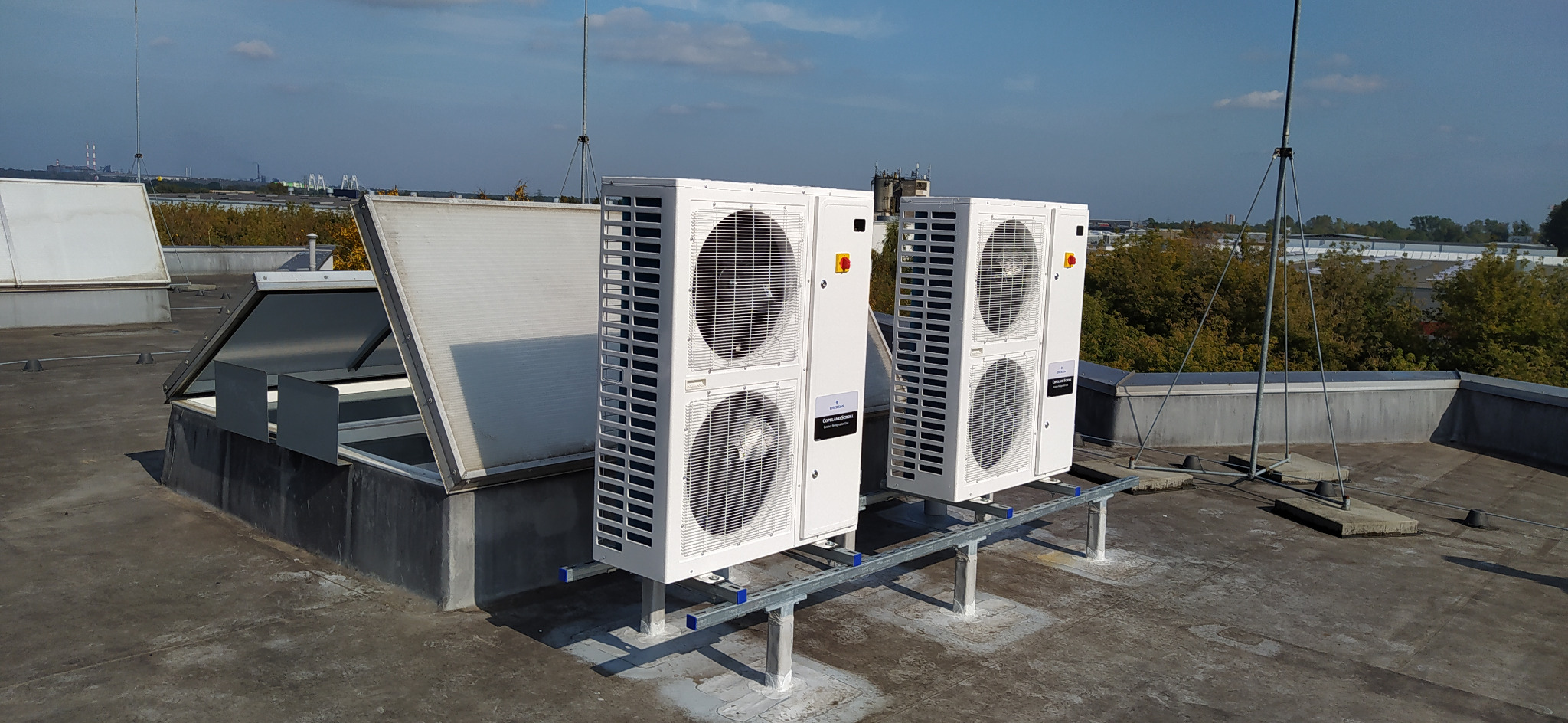 Dwa białe, bliźniacze urządzenia na dachu, prawdopodobnie klimatyzacyjne, zamontowane na metalowej konstrukcji, z widocznymi wentylatorami i elementami elektrycznymi, na tle miejskiego krajobrazu...