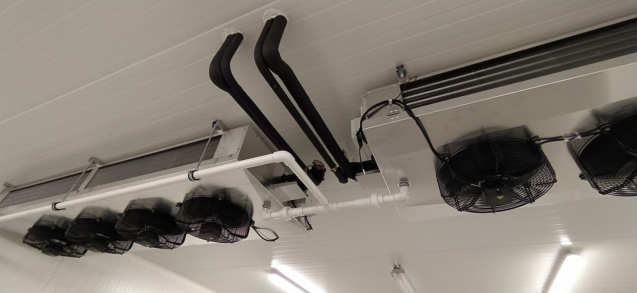 Widok od dołu na zamontowany system wentylacyjny z czarnymi rurami izolacyjnymi i białymi rurami PCV w pomieszczeniu o białych ścianach i suficie.