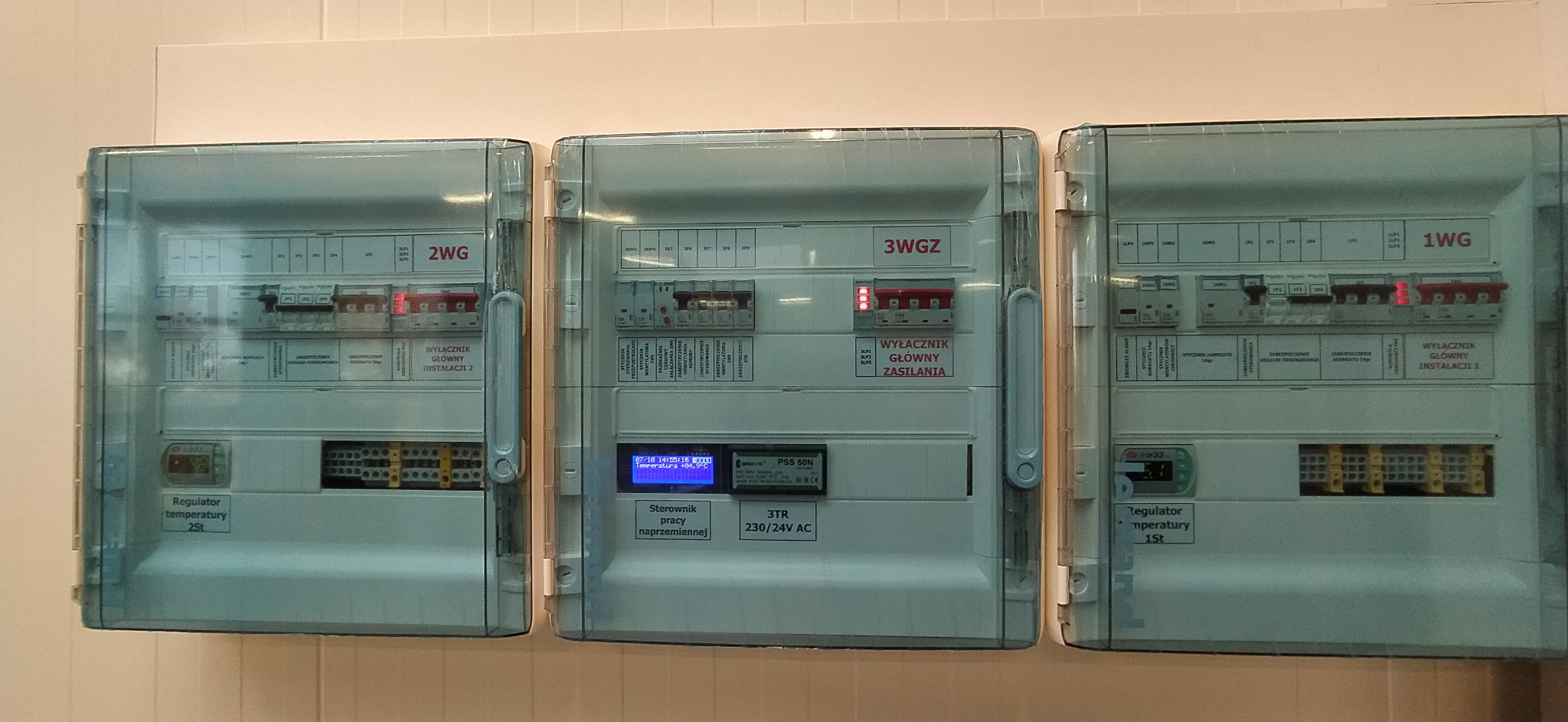 Trzy skrzynki sterownicze z wyłącznikami i regulatorami temperatury, oznaczone 1WG, 2WG i 3WGZ, zamontowane na białej ścianie, widoczne wyświetlacze i okablowanie.