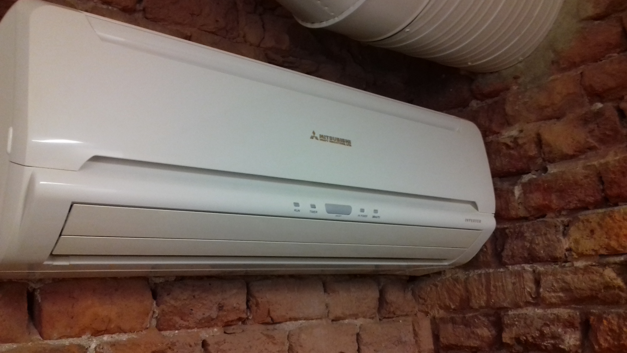 Biały klimatyzator Mitsubishi zamontowany na ścianie z czerwonej cegły, widoczny fragment rury wentylacyjnej pod sufitem.