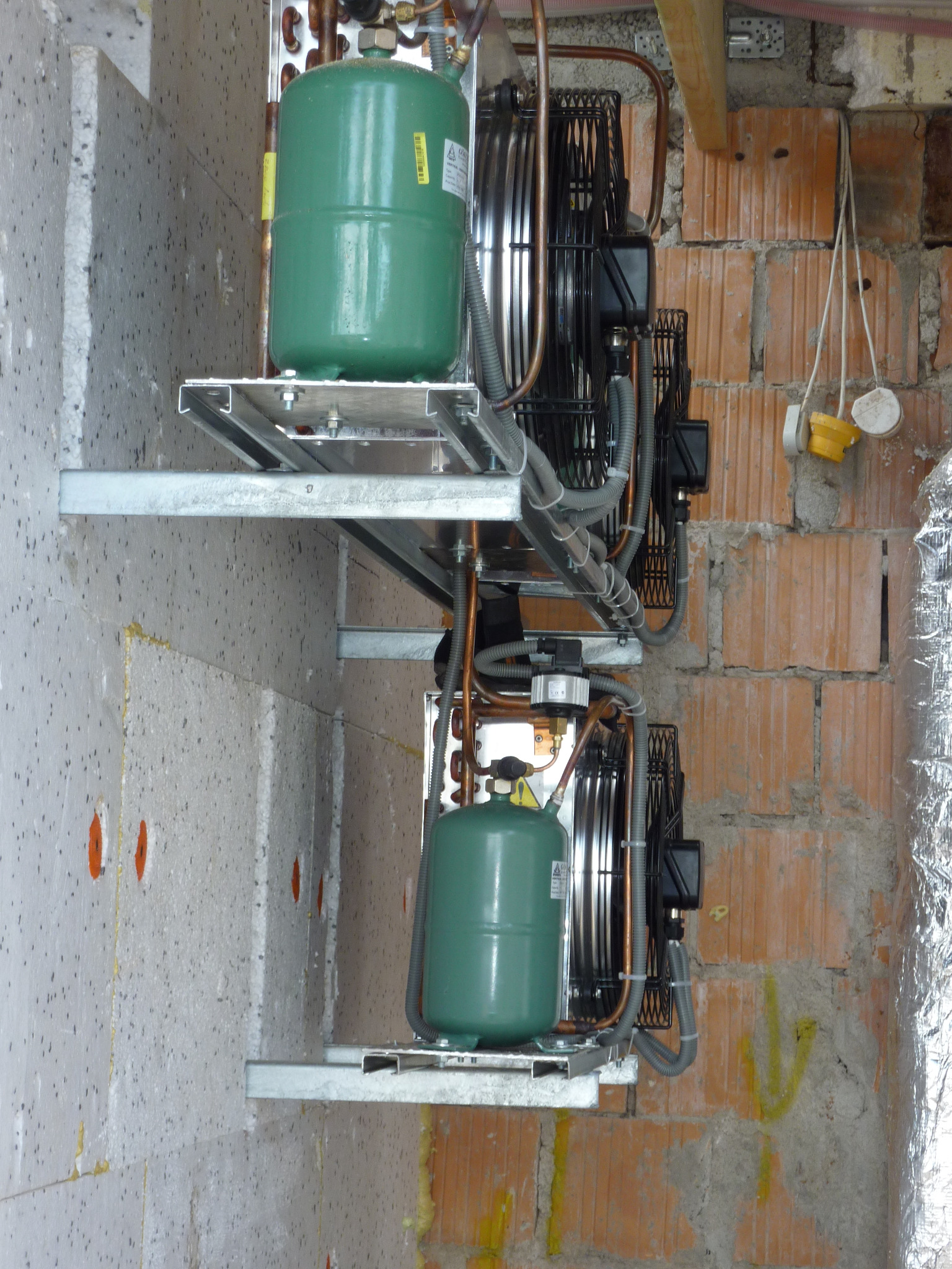 Dwa zielone zbiorniki na metalowych wspornikach, prawdopodobnie elementy systemu grzewczego, zamontowane na ścianie z izolacją styropianową i fragmentem ściany z czerwonej cegły. Widoczne przewody...