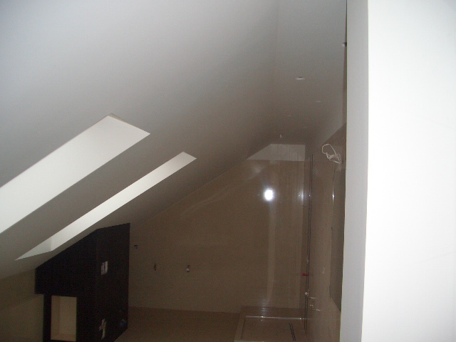 Surowe wnętrze poddasza ze skośnym sufitem, widoczne fragmenty instalacji elektrycznej i przygotowane ściany do malowania lub wykończenia, brak umeblowania.