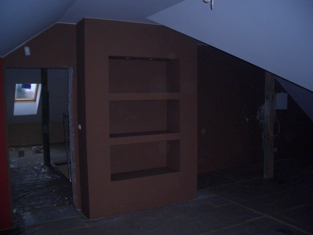 Poddasze w trakcie prac wykończeniowych: ściany pomalowane na brązowo, widoczna konstrukcja z płyt gipsowo-kartonowych z trzema wnękami, podłoga zabezpieczona folią, widoczne kable elektryczne...