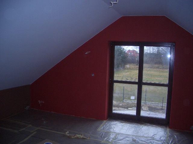 Pokój na poddaszu po malowaniu ścian na czerwono, zabezpieczona folią podłoga, widoczne ślady po gładzi szpachlowej na ścianach, okno balkonowe z widokiem na ogród.