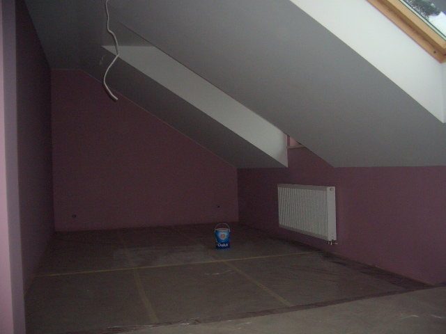Poddasze w trakcie prac wykończeniowych: ściany pomalowane na fioletowo, zabezpieczona podłoga, grzejnik na ścianie, widoczne płyty gipsowo-kartonowe na skosach sufitu oraz wiadro z farbą.