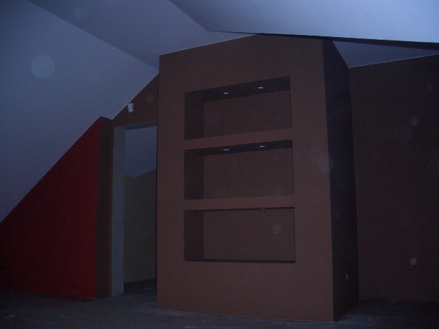Wnętrze w trakcie prac wykończeniowych: ściana z trzema wnękami oświetlonymi punktowo, czerwona ściana w kształcie trójkąta oraz otwór drzwiowy w szarym kolorze.