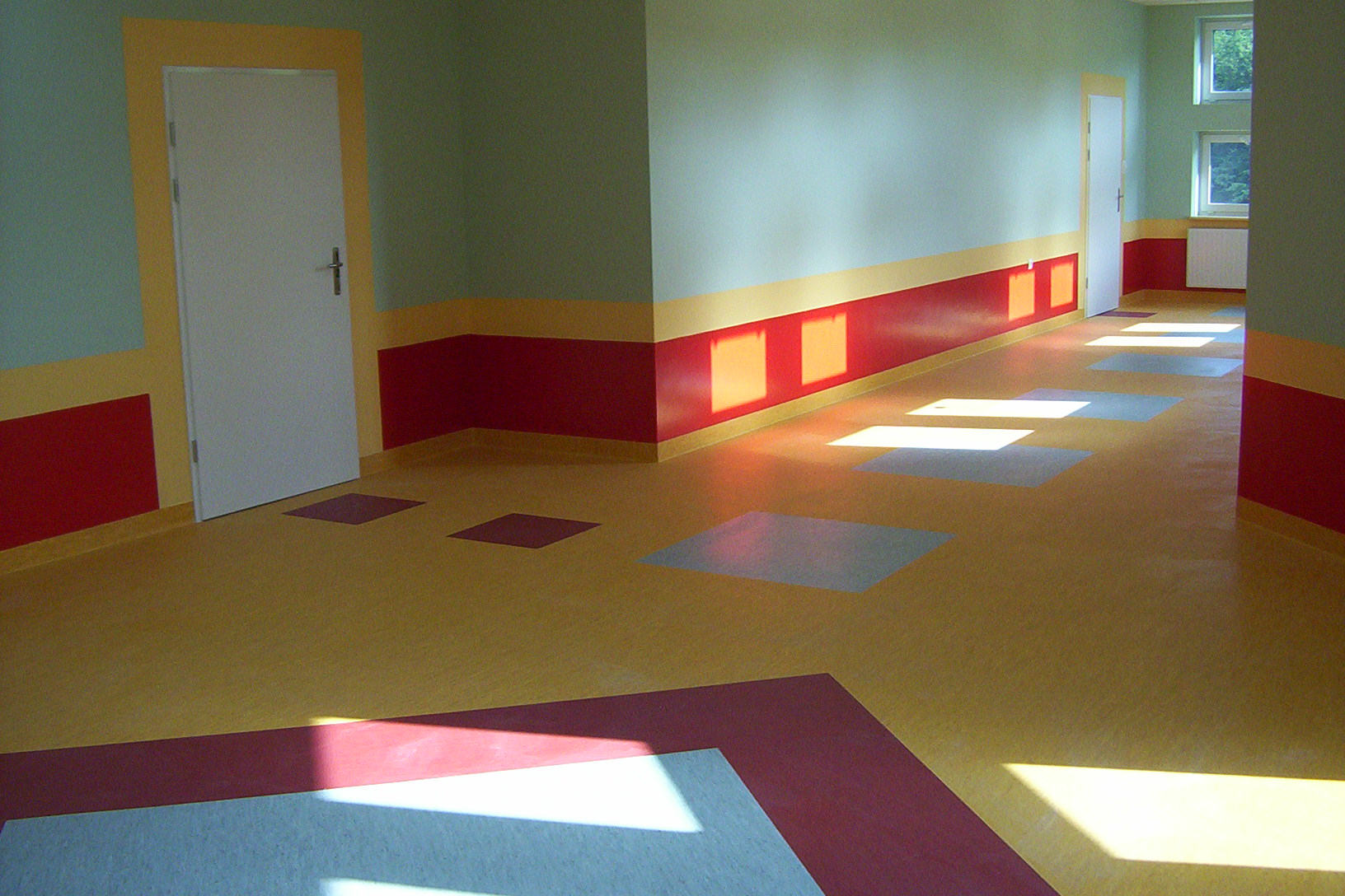 Korytarz z żółtą podłogą z wstawkami w kształcie kwadratów i rombów w kolorach czerwonym i szarym, ściany pomalowane na jasnozielono z czerwonym pasem na dole i żółtym paskiem pomiędzy, białe drzwi...