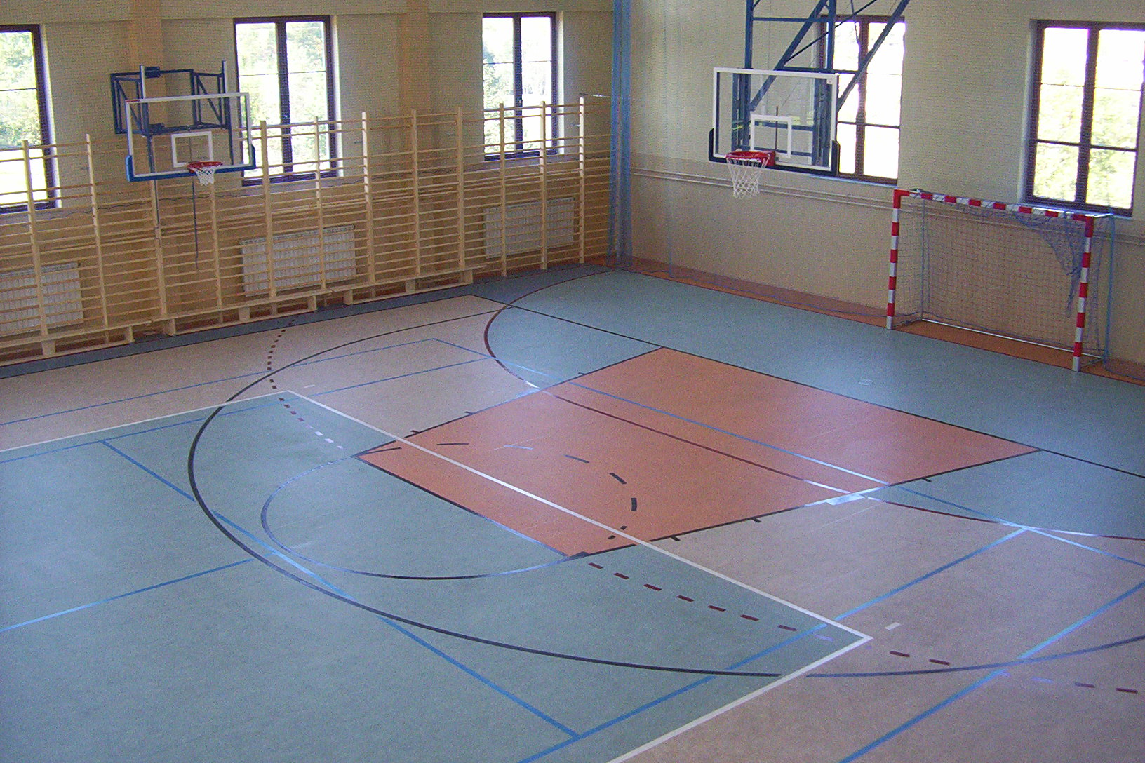 Widok z góry na salę gimnastyczną z parkietem w odcieniach szarości, zieleni i beżu, z liniami boisk do koszykówki i piłki ręcznej, z bramką do piłki ręcznej po prawej i koszami do koszykówki po...