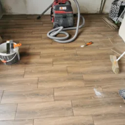 Układanie brązowych płytek podłogowych imitujących drewno, z widocznym wiadrem z zaprawą, szpachelką, odkurzaczem przemysłowym i szczotką, podczas prac wykończeniowych w pomieszczeniu.