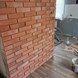 Ściana wykończona czerwoną cegłą klinkierową, widoczne niedociągnięcia w fugach, podłoga wyłożona panelami imitującymi drewno, w tle narzędzia i akcesoria remontowe.
