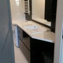 Nowoczesna łazienka z czarnymi szafkami pod umywalką z białym blatem, grzejnikiem drabinkowym i dużym lustrem w czarnej ramie.