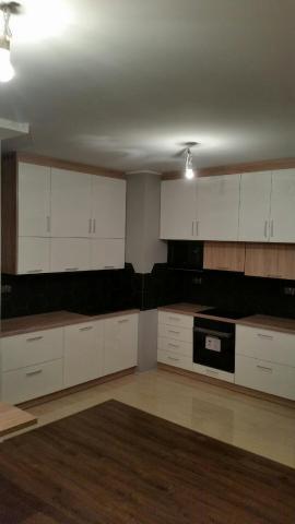 Nowoczesna kuchnia z białymi frontami szafek i drewnianymi blatami, czarny panel nad blatem, widok z perspektywy podłogi z paneli.