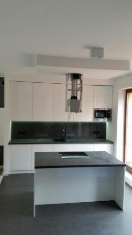 Nowoczesna kuchnia z białymi szafkami i czarnym blatem, wyspa kuchenna z płytą indukcyjną, oświetlenie LED pod szafkami, minimalistyczny okap.