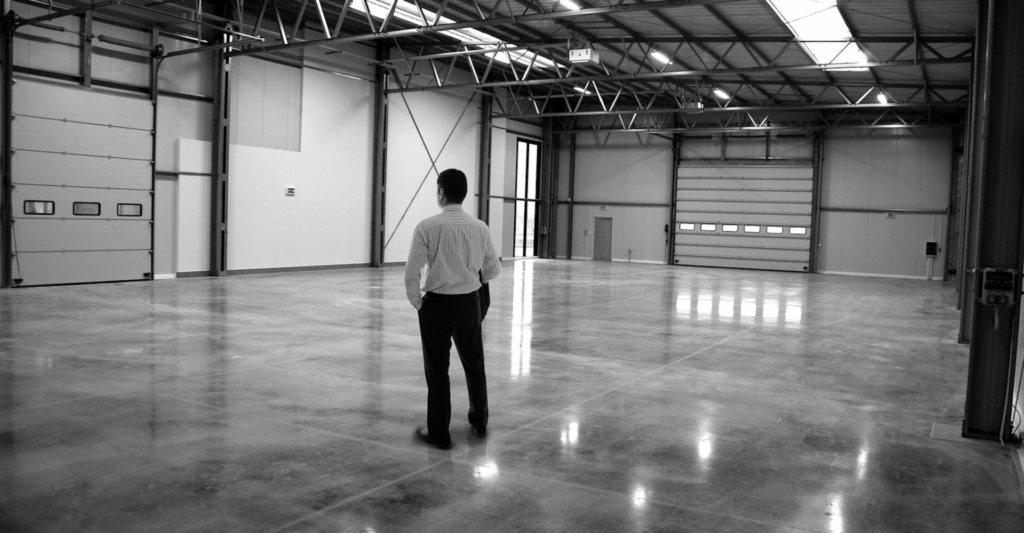 Czarno-białe zdjęcie mężczyzny stojącego w pustym, przestronnym magazynie w Komornikach z podłogą z polerowanego betonu i widocznymi bramami segmentowymi.
