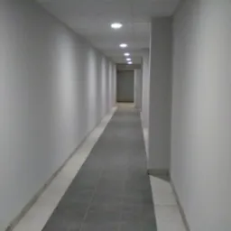 Długi korytarz z szarą podłogą i białymi ścianami, oświetlony rzędem okrągłych lamp sufitowych, z drzwiami na końcu.