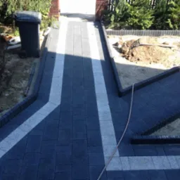 Ułożona, grafitowa kostka brukowa z białymi pasami, tworząca ścieżkę ogrodową. Widoczne obrzeża, przygotowane podłoże pod rabatę i kosz na śmieci w tle.