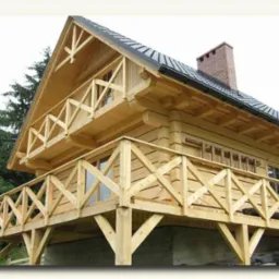 Jasny, dwukondygnacyjny dom z bali z balkonami, wykończony drewnianą balustradą, na betonowej podmurówce. Widoczny komin i fragment dachu z orynnowaniem.