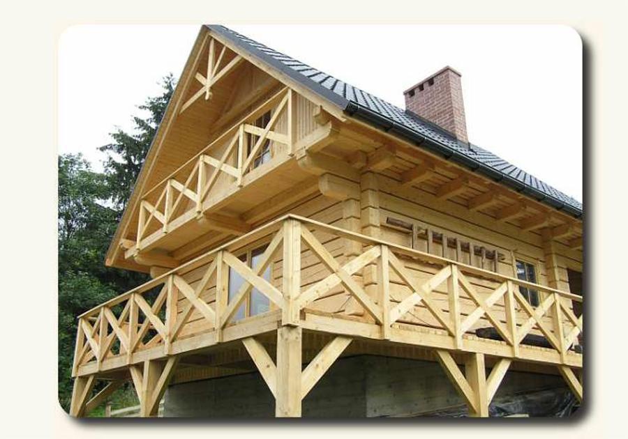 Jasny, dwukondygnacyjny dom z bali z balkonami, wykończony drewnianą balustradą, na betonowej podmurówce. Widoczny komin i fragment dachu z orynnowaniem.