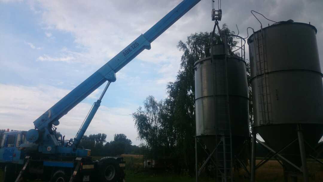 Niebieski dźwig AOK125 podnosi silos zbożowy na tle drzew i pochmurnego nieba.