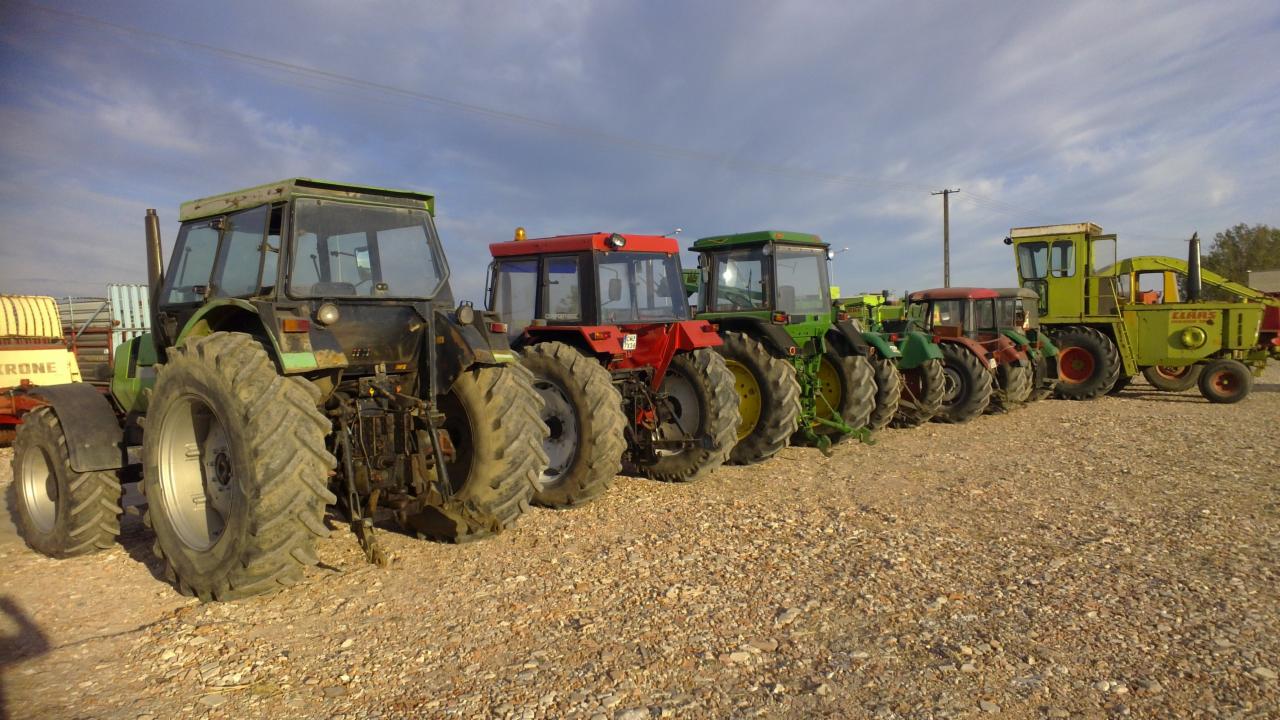Rząd używanych, różnokolorowych traktorów i kombajnów rolniczych, zaparkowanych na żwirowym placu pod błękitnym niebem z chmurami. Widok z boku, lekko z dołu.