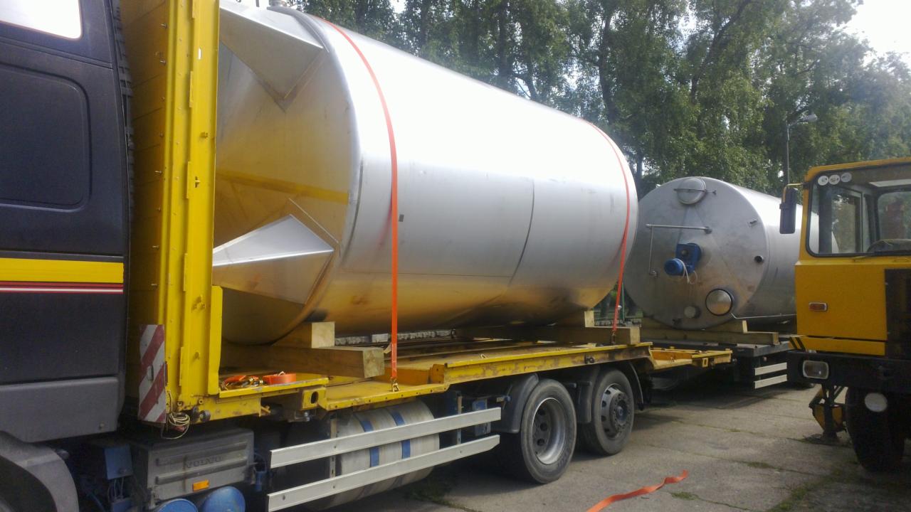 Transport dwóch dużych, srebrnych zbiorników na żółtej platformie ciężarówki z czarną kabiną. Jeden zbiornik ma stożkowe zakończenie. W tle drzewa.