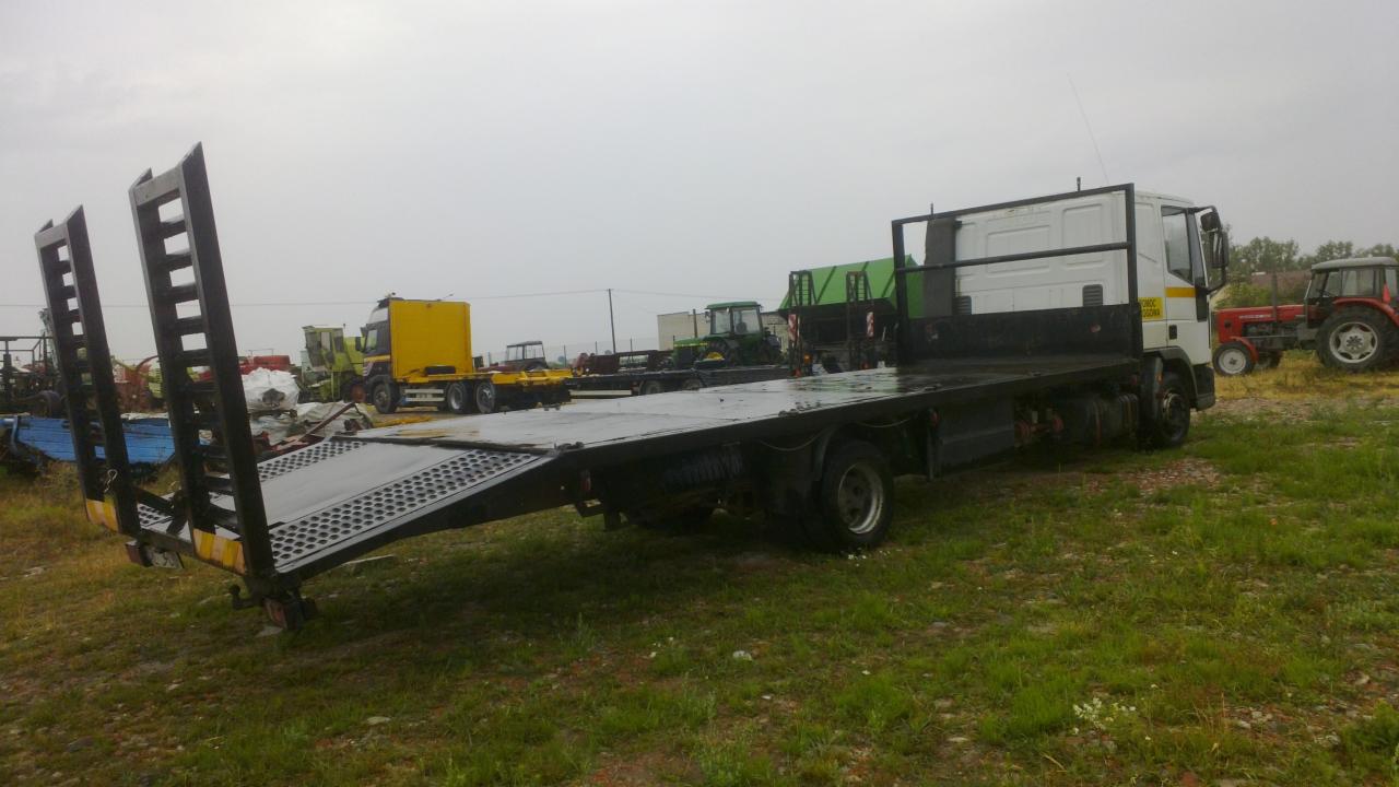 Ciężarówka z platformą załadunkową na trawiastym terenie, w tle inne pojazdy rolnicze, pochmurne niebo.