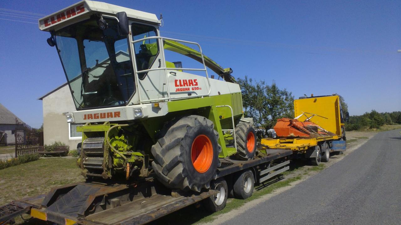 Kombajn Claas Jaguar 690SL na niskopodwoziowej platformie transportowej, z żółtą ciężarówką w tle, na tle błękitnego nieba i zieleni drzew.