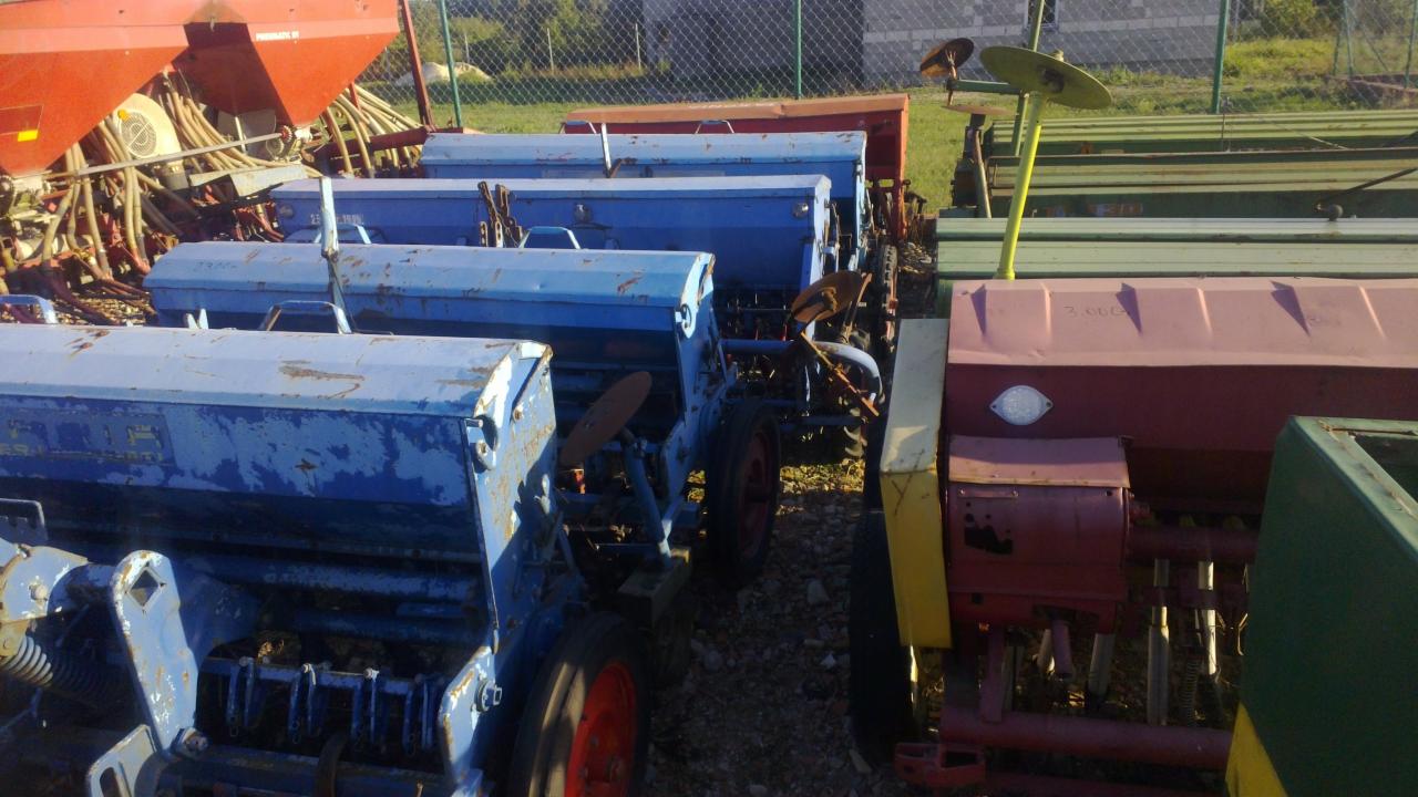 Rząd używanych, niebieskich siewników rolniczych na zewnątrz, z widocznymi śladami rdzy i zużycia, ustawionych obok siebie na żwirowym podłożu.