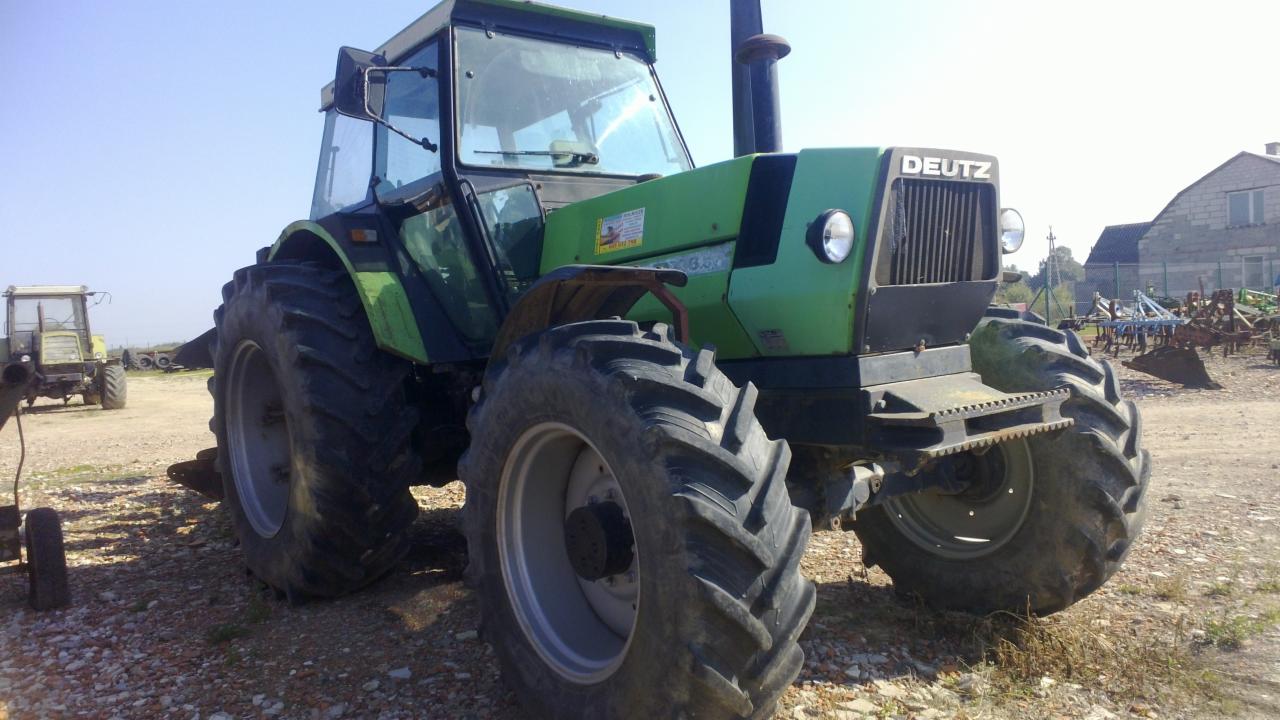 Zielony traktor Deutz na polnej drodze, z widocznymi dużymi oponami i kabiną. W tle budynek gospodarczy i inne maszyny rolnicze.