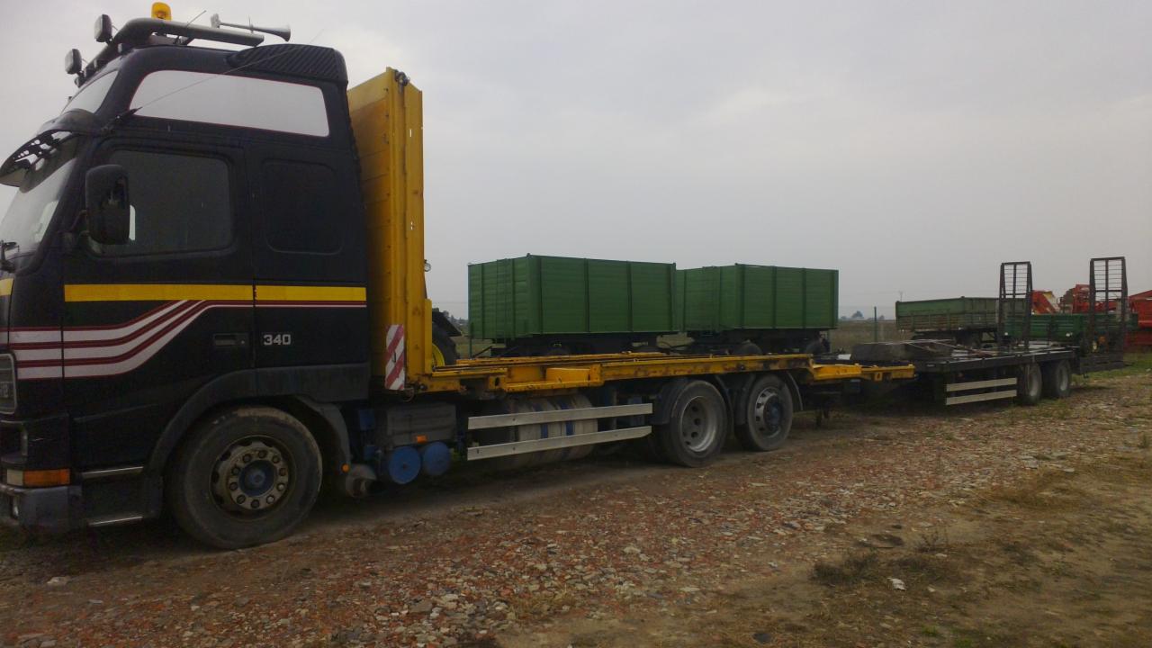 Czarno-żółty transport ciężarowy z zielonymi kontenerami na naczepie, widok z boku na żwirowym podłożu. Na dalszym planie sprzęt rolniczy.