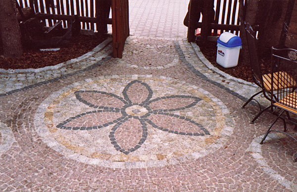 Ułożona z kostki brukowej ścieżka z motywem kwiatowym w odcieniach różu, szarości i beżu, prowadząca do drewnianej furtki, z widocznym koszem na śmieci i metalowym krzesłem w tle.