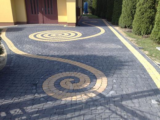 Ułożona kostka brukowa w kolorze szarym z żółtymi elementami tworzącymi spiralny wzór na podjeździe przed domem.