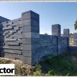 Nowoczesne ogrodzenie modułowe z szarej kostki łupanej, z widocznym fundamentem, w tle dom i drzewa.