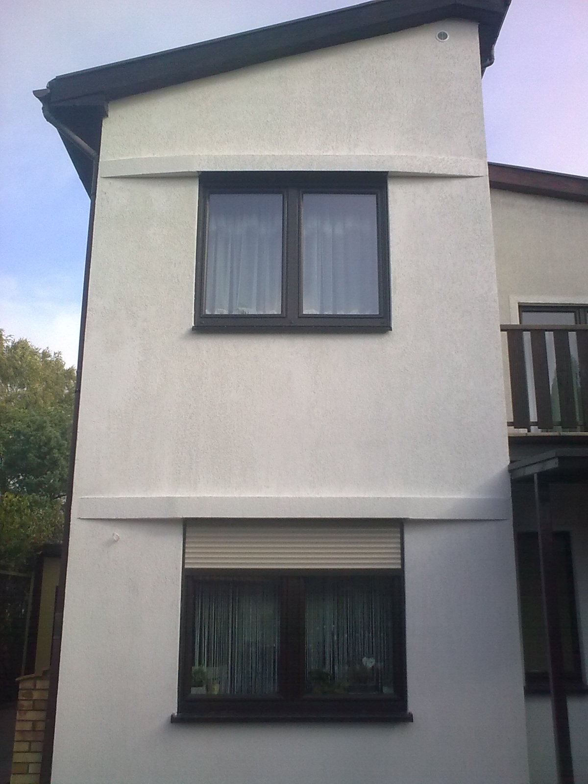 Elewacja dwukondygnacyjnego budynku z białym tynkiem, brązowymi oknami i roletami zewnętrznymi. Widoczne poziome pasy dekoracyjne na fasadzie.