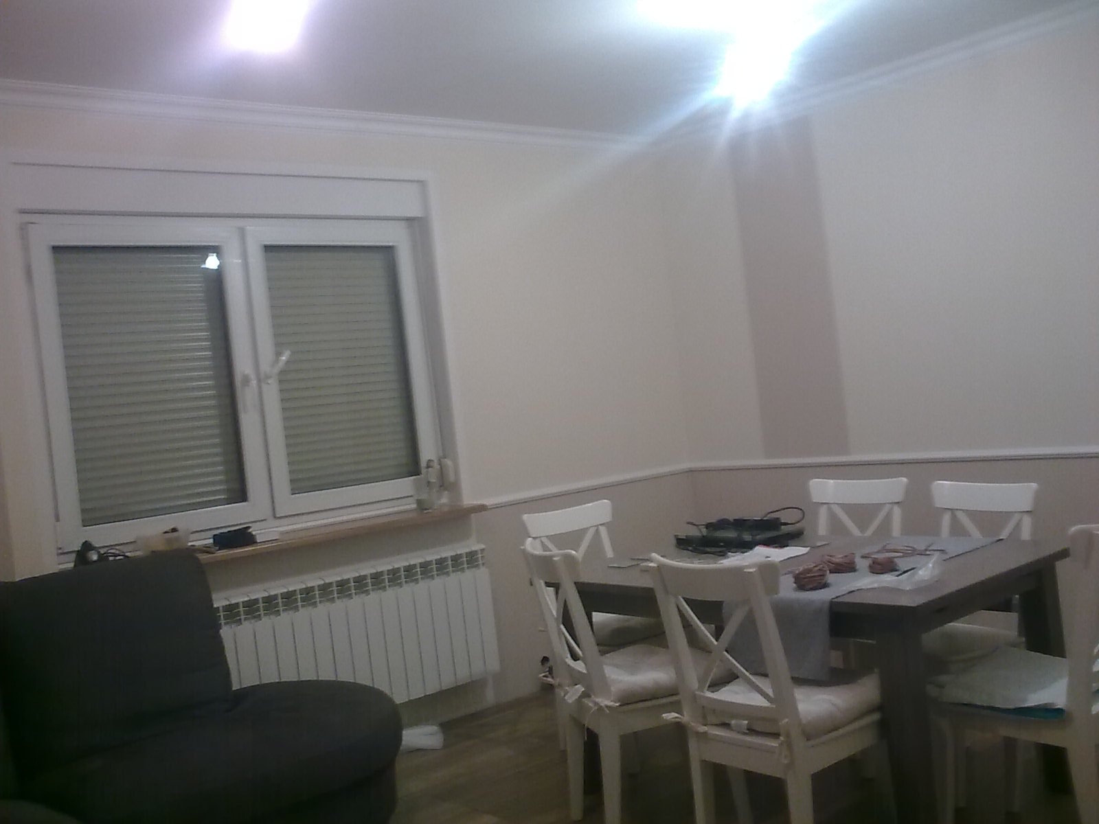 Jasne wnętrze pokoju po remoncie z białymi ścianami, oknem z żaluzjami, grzejnikiem, ciemnoszarą kanapą i drewnianym stołem z białymi krzesłami.