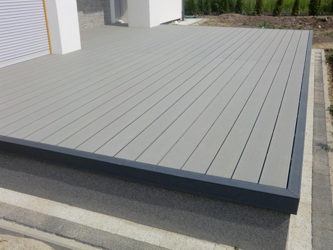 Szary taras kompozytowy przylegający do budynku, z widoczną strukturą desek i ciemną obwódką, na tle zieleni.