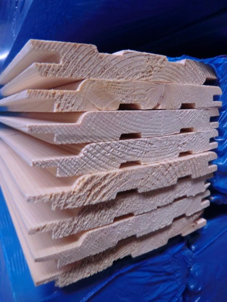 Stos jasnych, profilowanych desek z widocznym rysunkiem słojów drewna, zapakowanych w niebieską folię.