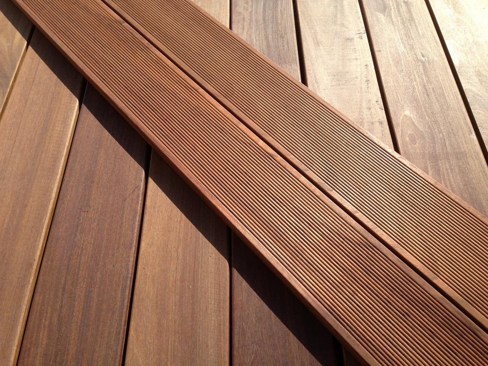 Dwie deski tarasowe z ryflowaną powierzchnią ułożone ukośnie na drewnianym podłożu o zróżnicowanym odcieniu brązu, oświetlone naturalnym światłem słonecznym.