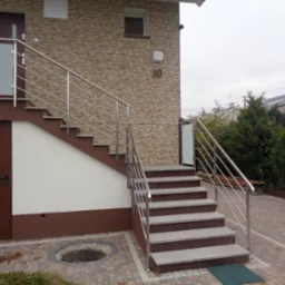 Zewnętrzne schody z szarego kamienia prowadzące do budynku z elewacją z cegły, zabezpieczone nowoczesną balustradą ze stali nierdzewnej i szkła, z numerem domu '10' widocznym na ścianie.